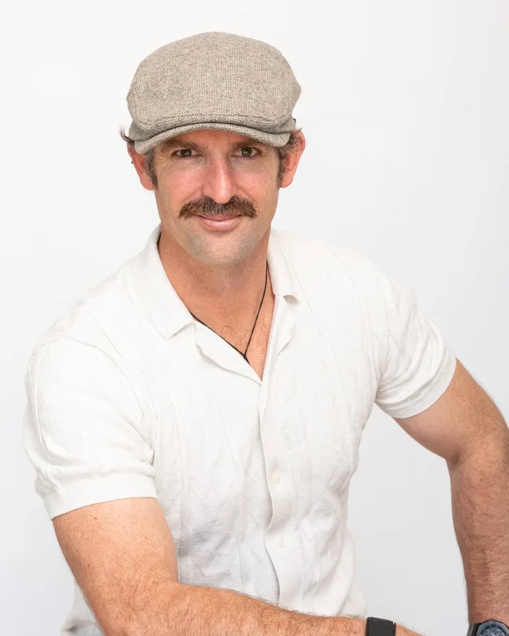Studio portrait of a man in a flat cap and a cream knitted shirt, seated, looking to camera.